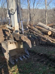An excavator is being used to remove logs from a wooded area.